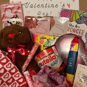 Valentine's Day care package image. The box has a cute "Happy Valentine's Day"sign. Inside, there is a variety of themed items, including candies, personal care items, socks, and a teddy bear. Everything is Valentine's Day colored.