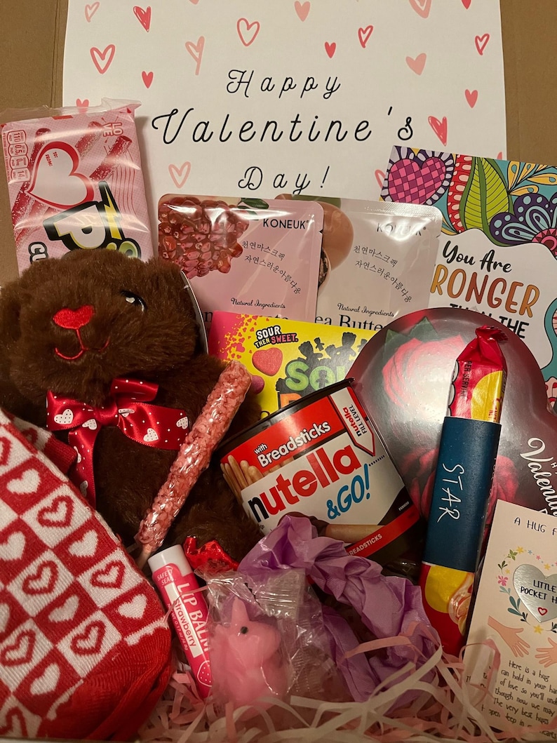 Valentine's Day care package image. The box has a cute "Happy Valentine's Day"sign. Inside, there is a variety of themed items, including candies, personal care items, socks, and a teddy bear. Everything is Valentine's Day colored.