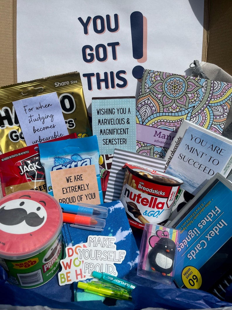 K&ouml;nnte beinhalten: Eine Geschenkbox gef&uuml;llt mit verschiedenen Artikeln f&uuml;r einen Studenten, darunter Snacks, Schreibwaren und motivierende Botschaften. Die Box enth&auml;lt eine T&uuml;te Schokoladen-Brezeln, eine T&uuml;te Erdnussbutter-Cracker, ein Glas Nutella, ein Pack Indexkarten, ein Notizbuch mit Mandala-Design, eine blau-wei&szlig; gestreifte Socke, eine Pinguin-Figur und mehrere motivierende Aufkleber.