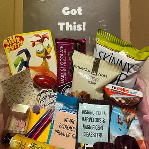 K&ouml;nnte beinhalten: Eine rosa und wei&szlig;e Geschenkbox gef&uuml;llt mit verschiedenen Artikeln, darunter Schokolade, Snacks, Schreibwaren und ein rotes Gummik&uuml;ken. Die Box hat eine Karte oben, auf der "You've Got This!" steht, und eine weitere Karte, auf der "Wishing you a marvelous & magnificent semester!" steht.