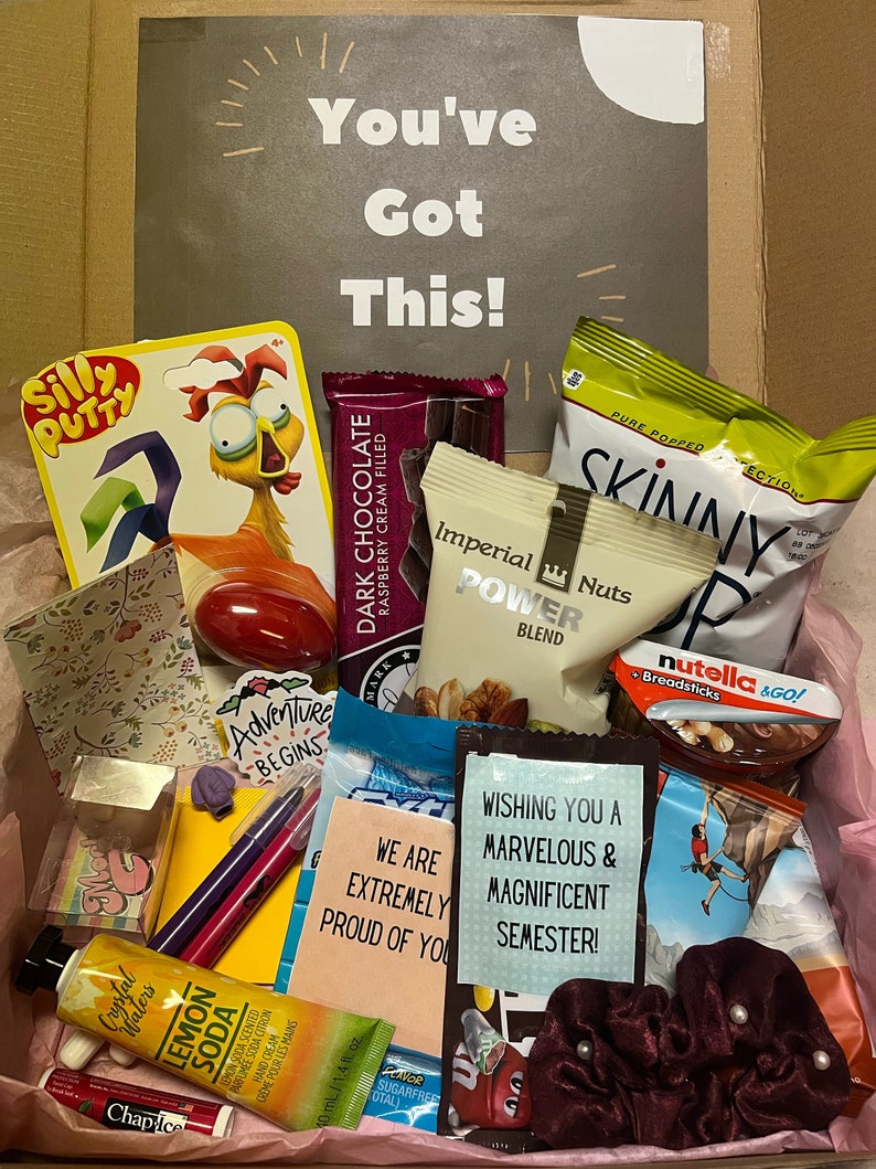 K&ouml;nnte beinhalten: Eine rosa und wei&szlig;e Geschenkbox gef&uuml;llt mit verschiedenen Artikeln, darunter Schokolade, Snacks, Schreibwaren und ein rotes Gummik&uuml;ken. Die Box hat eine Karte oben, auf der "You've Got This!" steht, und eine weitere Karte, auf der "Wishing you a marvelous & magnificent semester!" steht.