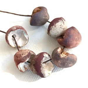 May include: A strand of seven irregular, cup-shaped ceramic beads strung on a thin brown cord. Each bead features a textured, matte exterior in shades of earthy brown, reddish-brown, and cream, with a smooth, hollowed-out white interior surface.