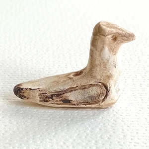 May include: A small, off-white ceramic bird figurine with a matte, textured surface. The bird is depicted in a seated position with dark brown painted accents along the wing and tail feathers, and a small circular hole on its back.