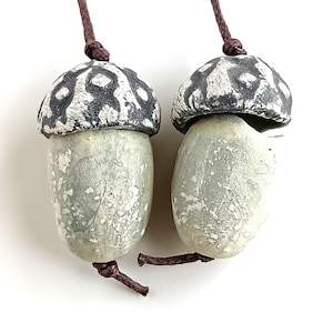 Pode incluir: Dois pingentes em forma de bolota com uma aparência rústica e artesanal. As tampas das bolotas são cinza escuro com um padrão branco, enquanto os corpos são verde pálido com uma superfície texturizada. Cada pingente é suspenso por um cordão marrom, amarrado na parte superior e inferior.