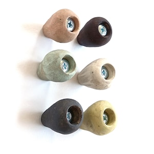 Könnte beinhalten: Sechs Klettergriffe in verschiedenen Farben, darunter Beige, Braun, Grün und Gelb. Jeder Griff hat eine abgerundete Form mit einer Aussparung für eine Schraube. Die Griffe sind für das Klettern und Bouldern konzipiert.