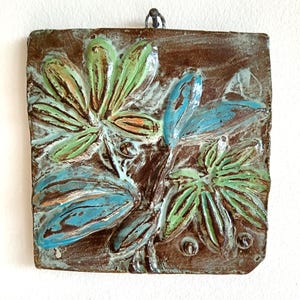Könnte beinhalten: Eine quadratische Wandplatte aus Keramik mit einem Reliefdesign aus stilisierten Blumen und Blättern. Das Stück hat eine dunkelbraune Grundglasur mit handgemalten Akzenten in Hellgrün und leuchtendem Blau sowie eine glänzende, strukturierte Oberfläche.