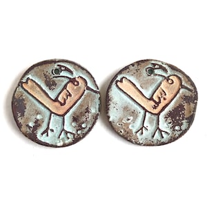 Puede incluir: Dos colgantes redondos de cerámica con diseño de pájaro. Los colgantes tienen un fondo azul claro y un borde marrón. El pájaro es de color canela con detalles negros. Cada colgante tiene un pequeño agujero en la parte superior.