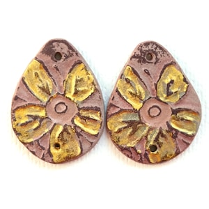 May include: Two teardrop-shaped pendants made of matte mauve clay, each featuring a central circular motif surrounded by five gold-painted petals. Each pendant has a small circular hole at the top for suspension and a textured, earthy surface finish.