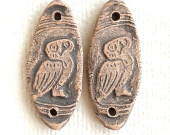 May include: Two oval-shaped clay beads featuring a relief design of an owl. Each bead has a textured, earthy brown surface with dark gray recessed areas and two circular holes, one at the top and one at the bottom, for threading purposes.