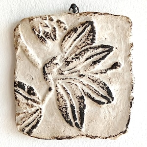 May include: A square, off-white wall plaque featuring a raised, dark brown relief of a botanical floral design. The surface has a textured, matte finish with uneven edges and a small black metal loop at the top centre for hanging purposes.