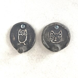 Puede incluir: Dos perchas de pared de cerámica gris oscuro. Una percha presenta un diseño de búho blanco y la otra una cara de gato blanco. Cada percha tiene una forma pequeña y redonda con un tornillo para el montaje.