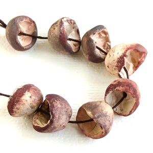 Peut inclure: Un collier composé de huit perles en céramique creuses en forme de coupelle, enfilées sur un cordon marron foncé. Chaque perle présente un extérieur texturé et tacheté dans des tons brun-rouge et crème, avec un intérieur blanc cassé lisse.