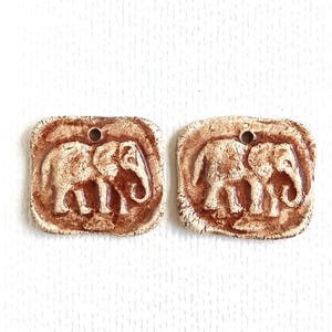 Puede incluir: Dos colgantes cuadrados de cerámica con esquinas redondeadas, cada uno con un relieve de elefante. Las piezas tienen un acabado agrietado y desgastado en tonos marrón terracota y blanco roto, con un pequeño orificio superior para colgar.