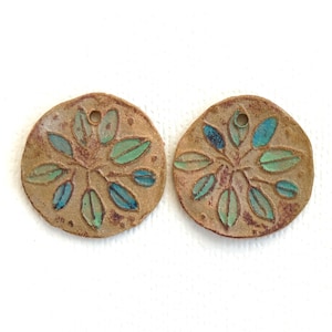 May include: Two circular tan clay pendants featuring a debossed leaf pattern. Each leaf is filled with shades of teal, blue, and green glaze. The pendants have a rough, textured surface and a single small hole at the top for hanging or attachment.