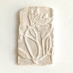 Puede incluir: Una placa de pared rectangular de arcilla color hueso con un diseño en relieve de una flor con pétalos y hojas alargadas. La superficie tiene un acabado mate y texturizado con bordes irregulares formados a mano y un aspecto natural.