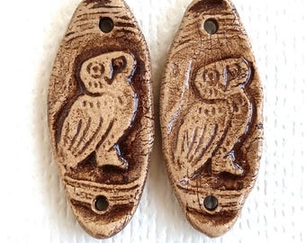 May include: Two oval-shaped ceramic beads featuring a relief design of an owl. The beads have a light tan base color with dark brown glazed accents in the recessed areas and a crackled texture. Each bead has two holes for threading purposes.