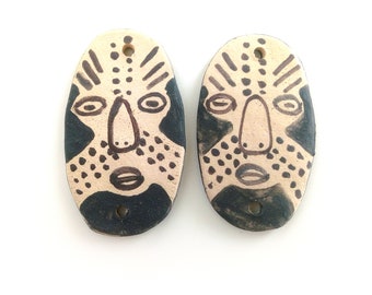 May include: Two ceramic oval-shaped earrings with a black and white painted design of a face. The face has a black outline and brown details.