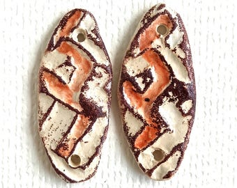 May include: Two elongated oval ceramic beads with a textured, off-white base glaze. Each bead features a hand-painted pattern of dark brown and burnt orange geometric shapes and lines. There is a single circular hole at the top and bottom of each.