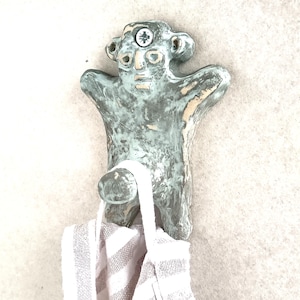 May include: A handmade ceramic wall hook in the shape of a stylized figure with outstretched arms. The hook is a mottled blue-green color with a screw in the head. A striped towel hangs from the hook.