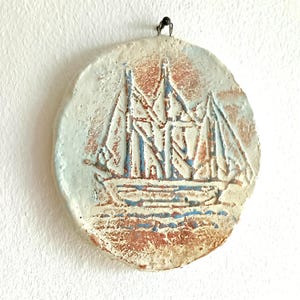 May include: A circular wall plaque featuring a textured, relief-style carving of a sailing ship. The piece is finished in a distressed palette of off-white, terracotta orange, and pale blue, with a small metal loop at the top for hanging.