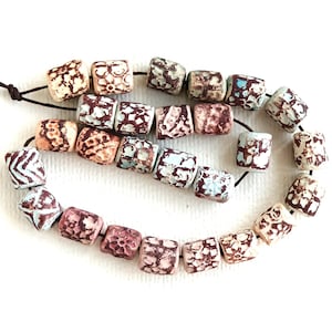 May include: A strand of cylindrical beads strung on a dark brown cord. Each bead features a textured, matte surface with a distressed finish, displaying intricate patterns in shades of cream, light blue, and deep reddish-brown throughout the set.