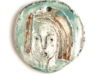 May include: A handmade ceramic pendant with a stylized face, brown hair, and black outlines. It has a light blue border and a hole for hanging. The ceramic has a textured, rustic appearance.