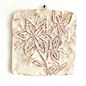 May include: A square, off-white textured wall plaque featuring a relief carving of a large flower and leaves. The recessed areas of the floral design are filled with a reddish-brown pigment, creating a contrast against the light, uneven surface.