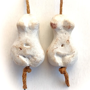 May include: Two small, off-white ceramic figurines of the female form, each suspended by a brown cord. The figurines have a rough texture with subtle brown markings. The cords are knotted at the bottom.