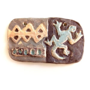May include: A ceramic tile with a brown, blue, and yellow glaze. The tile features a geometric pattern on the left side and a stylized lizard on the right side.