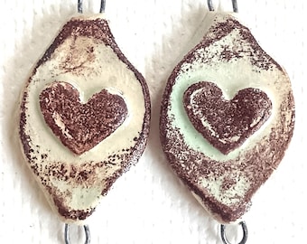 May include: Two diamond-shaped ceramic pendants featuring a raised brown heart in the center. The pendants have a cream-colored base with brown speckled edges and a metal loop at the top and bottom for attachment to jewelry or other items.