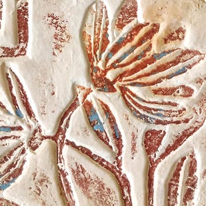 May include: A textured off-white surface featuring a relief carving of floral elements. The carved design is highlighted with distressed reddish-brown and faded blue paint, creating a weathered appearance with deep grooves and raised edges.