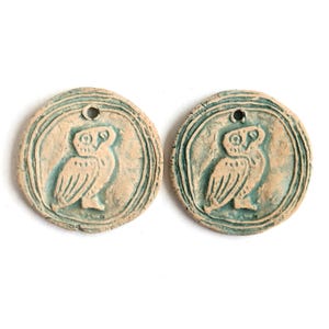 Könnte beinhalten: Zwei runde, hellfarbene Keramikanhänger mit einem türkisfarbenen Eulenmotiv. Jeder Anhänger hat oben ein kleines Loch zum Auffädeln. Die Eule ist innerhalb eines kreisförmigen Randes zentriert, und die Anhänger haben ein rustikales, handgemachtes Aussehen.