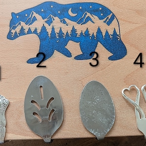 May include: Four silver serving utensils are displayed on a wooden cutting board with a blue bear silhouette design. The utensils include a decorative spoon, a slotted server, a solid server, and a fork with heart-shaped tines.