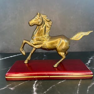 May include: A brass-colored horse figurine in a prancing pose, featuring a detailed mane and tail. The horse is mounted on a rectangular, dark reddish-brown wooden base with a thin gold-colored trim line running along the side of the base.