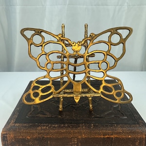 Puede incluir: Un sujetalibros de metal dorado en forma de mariposa con un diseño intrincado. El sujetalibros está sobre un pequeño caballete de madera.