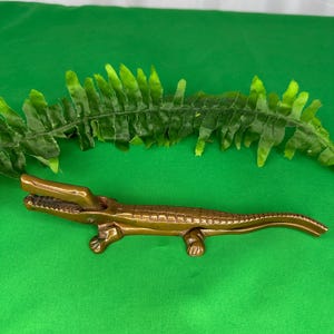 May include: A brass alligator-shaped nutcracker with a hinged jaw mechanism. The metal figure features a textured, scaled back, four short legs, and a long tail, with the upper jaw positioned in an open state to reveal detailed teeth.