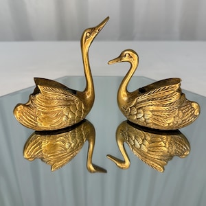 Peut inclure: Deux figurines de cygnes en laiton avec des textures de plumes détaillées sur leurs ailes. Un cygne a un long cou droit, tandis que l'autre a un cou plus court et courbé. Les deux figurines présentent des centres creux et sont en laiton doré.