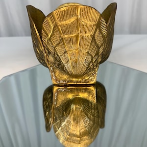 May include: A gold-colored metal bowl shaped like a large seashell. The exterior features a textured, ribbed pattern that mimics the natural ridges of a scallop shell. The bowl has a wide, open top with curved edges and a reflective, metallic finish.