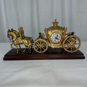 Puede incluir: Un reloj de carruaje dorado con dos caballos y jinetes tirando de un coche detallado. La esfera del reloj está integrada en el lateral del carruaje, que está rematado con un adorno de corona. La pieza está montada sobre una base de madera oscura.