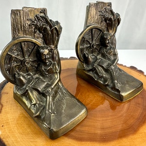 Puede incluir: Un par de sujetalibros de bronce que representan una escena rústica con un hombre sentado junto a una rueda de carro y un manojo de trigo. Los sujetalibros son detallados y tienen un aspecto vintage.