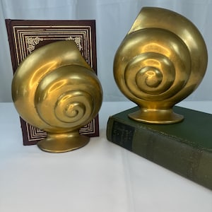 Puede incluir: Dos sujetalibros dorados en forma de concha espiral. Los sujetalibros son de metal y tienen un acabado brillante. Un sujetalibros está a la izquierda de un libro granate, y el otro a la derecha de un libro verde.