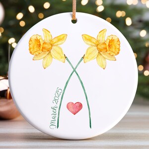 May include: White ceramic ornament with two yellow daffodils, green stems, and a small red heart. The text "March 2023" is written on the ornament.