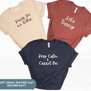 May include: Three t-shirts with different vegetable-themed puns. The first is a soft cream t-shirt with the text "Daisy See me Rollin". The second is a heather clay t-shirt with the text "Let's Turnip". The third is a heather navy t-shirt with the text "Keep Calm & Carrot On".