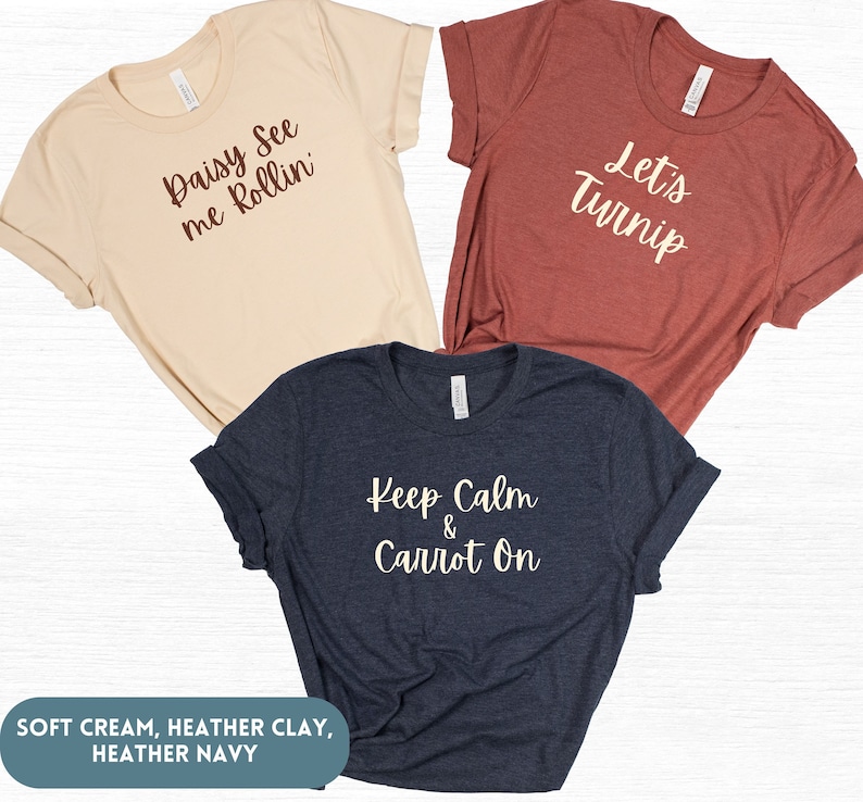 May include: Three t-shirts with different vegetable-themed puns. The first is a soft cream t-shirt with the text "Daisy See me Rollin". The second is a heather clay t-shirt with the text "Let's Turnip". The third is a heather navy t-shirt with the text "Keep Calm & Carrot On".