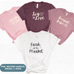 May include: Four t-shirts with different designs and colors. The shirts are pink, heather maroon, orchid, and white. The designs include "garden gal", "Soy in Love", "Bouquet Babe", and "Fresh off the Market".