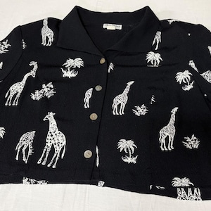 Op de afbeelding: Een zwart overhemd met korte mouwen en knoopsluiting, voorzien van een herhalende witte print van giraffen en palmbomen. Het kledingstuk heeft een klassieke kraag, een knoopsluiting aan de voorzijde met lichte knopen en een rechte zoom.