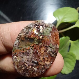 Pode incluir: Uma pedra preciosa oval com uma superfície texturizada. A pedra apresenta uma mistura de cores, incluindo tons de castanho, verde e branco, criando uma aparência natural e terrosa. A pedra preciosa é segurada entre os dedos, com folhagem verde no fundo.