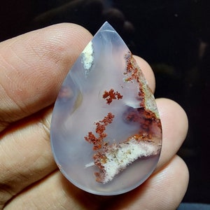 Peut inclure: Un cabochon d'agate en forme de larme avec une base blanche translucide et des inclusions dendritiques rouges et brunes.