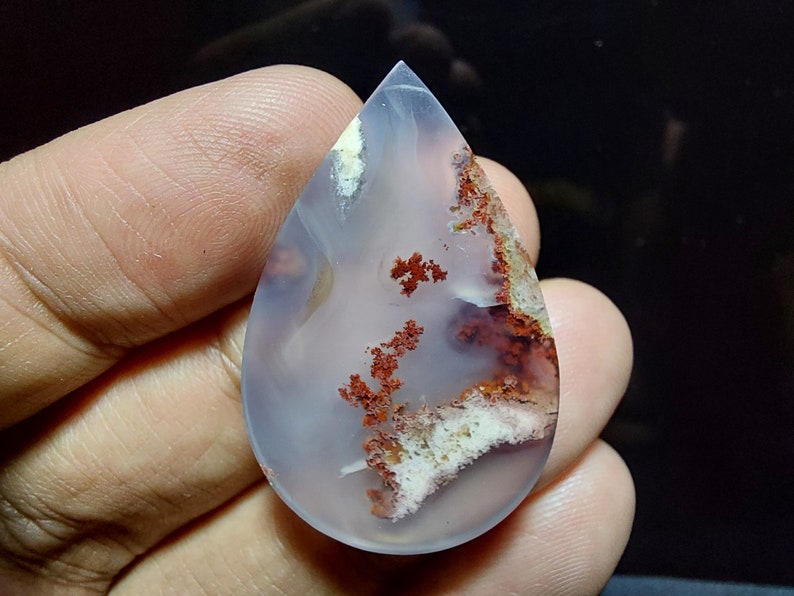 Peut inclure: Un cabochon d'agate en forme de larme avec une base blanche translucide et des inclusions dendritiques rouges et brunes.