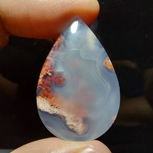 Peut inclure: Un cabochon d'agate en forme de larme avec un motif de couleur blanc, gris et rouge. L'agate a une apparence translucide avec une surface lisse et polie.
